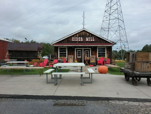 A rustic cider mill with outdoor seating, pumpkins, and a power tower in the background on a cloudy day.