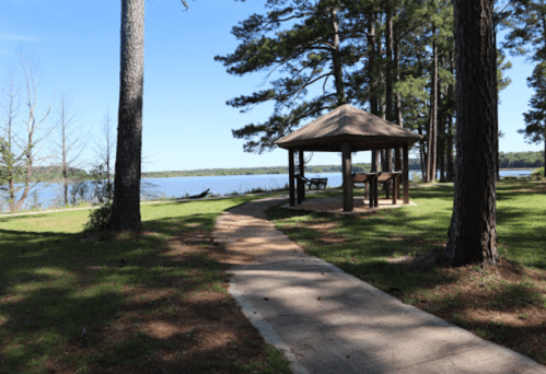 A serene lakeside scene featuring a path leading to a gazebo surrounded by tall trees and clear blue skies.