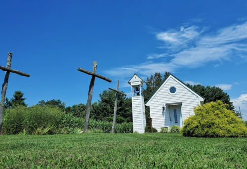 A small white church with a bell tower, surrounded by green grass and wooden crosses under a clear blue sky.