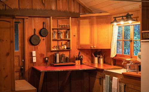 Cozy wooden kitchen with shelves, pots, and a red countertop, featuring warm lighting and rustic decor.