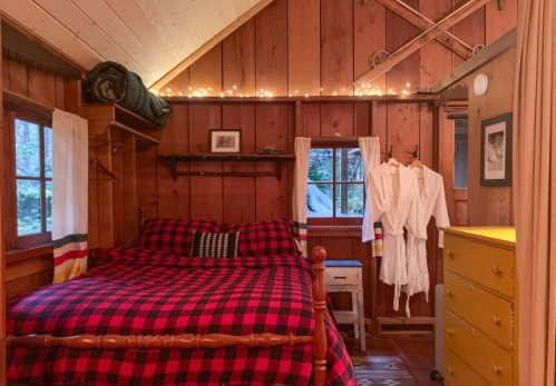 Cozy cabin interior with a plaid bedspread, wooden walls, and warm lighting, featuring a dresser and bathrobes.