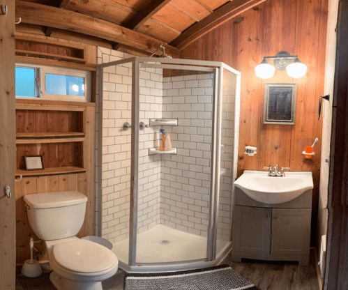 Cozy bathroom featuring a shower, toilet, sink, wooden walls, and shelves for storage.