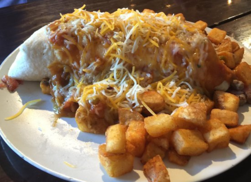 A large burrito topped with melted cheese and sauce, served with crispy potato cubes on a white plate.
