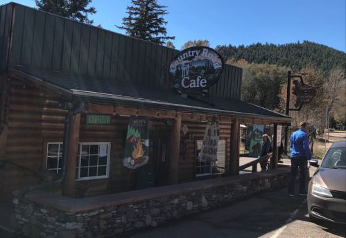 A rustic log cabin cafe with a sign reading "Country Rose Cafe" and a person standing outside on a sunny day.