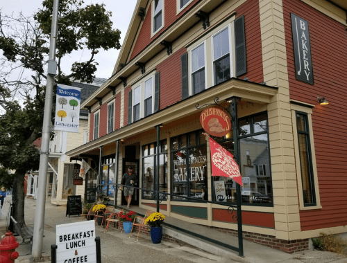 A charming bakery with a red and cream exterior, featuring outdoor seating and colorful flower pots.