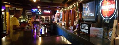 A dimly lit bar with a long counter, festive lights, and a TV showing a sports game. Two patrons are at the bar.