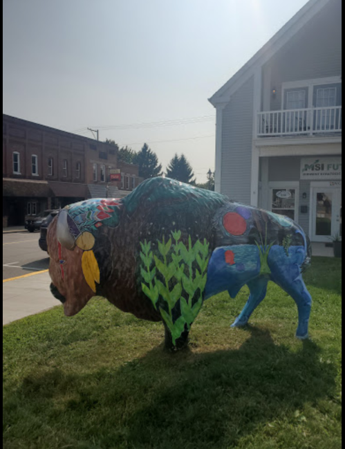 A colorful painted buffalo sculpture stands on grass near a building, with vibrant floral and abstract designs.