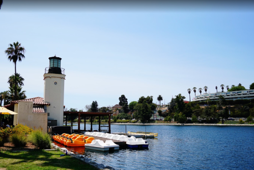 A scenic lakeside view featuring a lighthouse, colorful paddle boats, and palm trees under a clear blue sky.