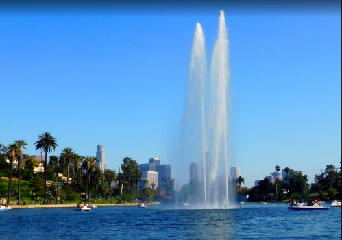 A fountain sprays water into the sky on a lake, with palm trees and city skyline in the background under a clear blue sky.