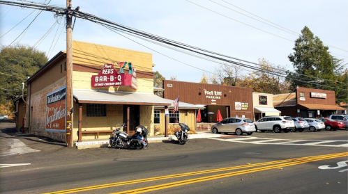 A street view of a barbecue restaurant and nearby shops, with parked cars and motorcycles outside.