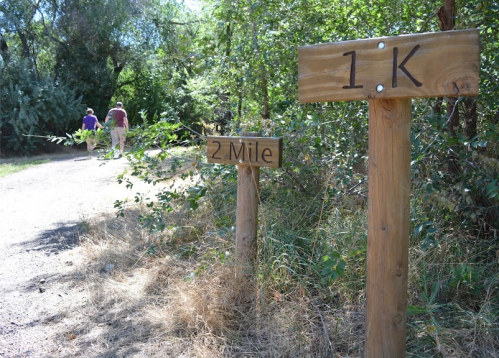 Two wooden trail signs indicate distances: "1 K" and "2 Mile," with a path and two people walking in the background.
