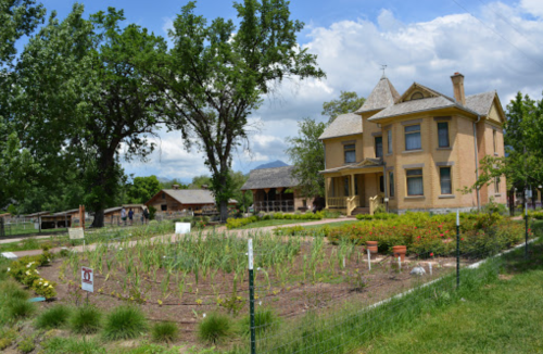 A historic yellow house surrounded by a garden with green plants and trees under a partly cloudy sky.