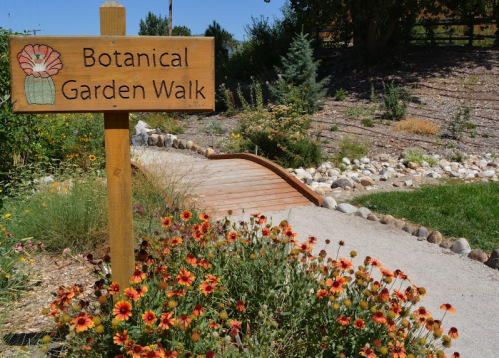 A wooden sign reads "Botanical Garden Walk" beside a flower-filled pathway in a lush garden setting.