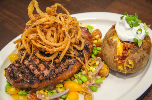 A grilled steak topped with crispy onions, served with a baked potato and a colorful vegetable medley.