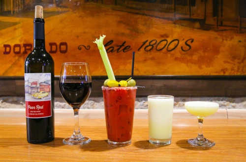A selection of drinks on a wooden table: a bottle of red wine, a glass of red wine, a Bloody Mary with celery, and a margarita.