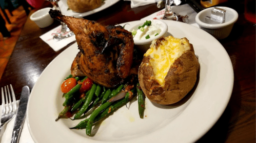 A plate featuring grilled chicken, green beans, cherry tomatoes, a baked potato, and a side of sour cream.