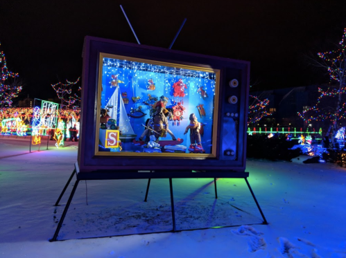 A large, colorful TV display with holiday decorations and figures, surrounded by festive lights in a snowy setting.