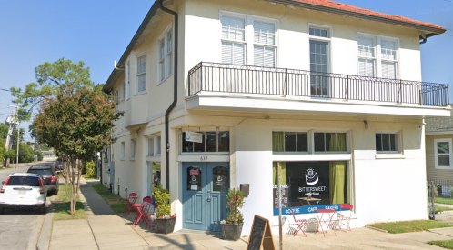 A charming café with outdoor seating, located in a two-story white building on a sunny street.