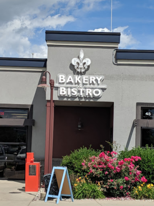 Sign for a bakery and bistro with a decorative fleur-de-lis, surrounded by flowers and a sidewalk chalkboard.
