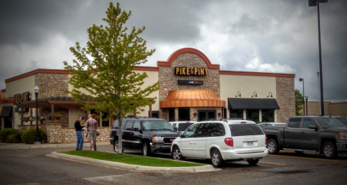 Exterior of Pike & Pin restaurant with parked cars and people chatting outside under a cloudy sky.