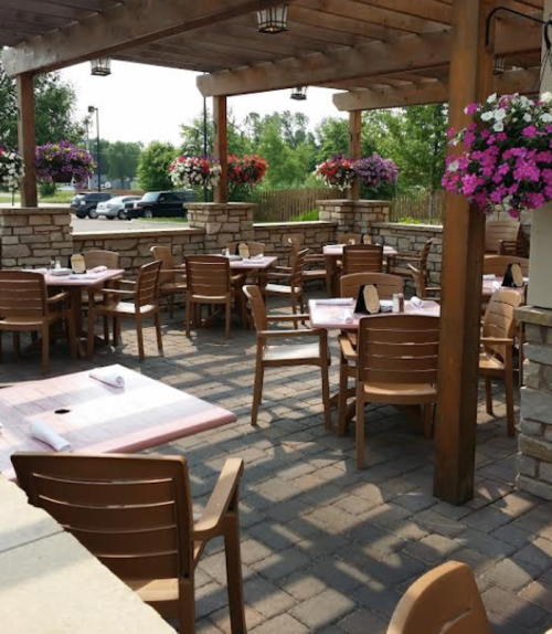 Outdoor dining area with wooden tables and chairs, surrounded by colorful flower pots and shaded by a wooden pergola.
