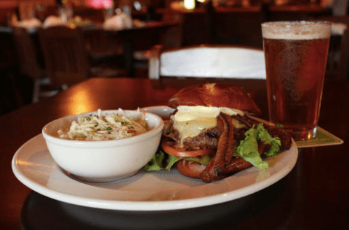 A plate with a burger topped with cheese and bacon, a side of coleslaw, and a glass of beer on a table.