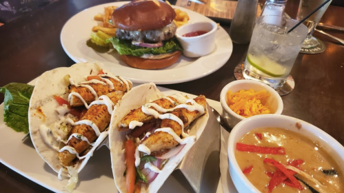 A plate with fish tacos, a burger with fries, and a bowl of soup, alongside a cocktail on a wooden table.