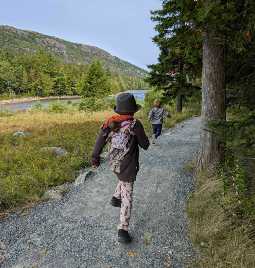 Two children walking along a gravel path by a river, surrounded by trees and mountains in the background.