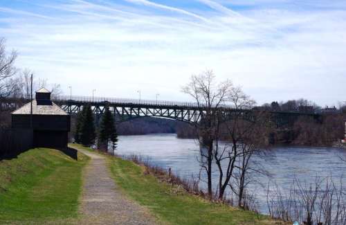 A scenic view of a river with a green bridge in the background, lined by trees and a path along the water's edge.