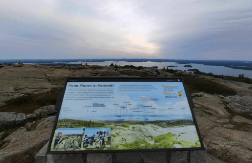 A scenic view from a mountain summit with a sign titled "From Shores to Summits" and a lake in the background.