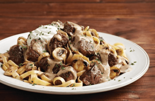 A plate of fettuccine topped with beef, mushrooms, creamy sauce, and a dollop of sour cream, garnished with herbs.
