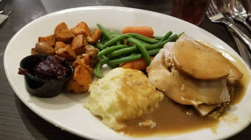 A plate of turkey with gravy, mashed potatoes, green beans, carrots, sweet potatoes, and a side of cranberry sauce.