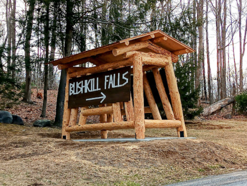 Wooden sign for Bushkill Falls with an arrow pointing right, surrounded by trees and grass.