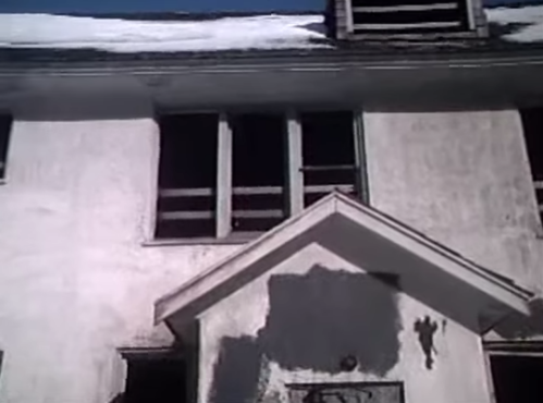 Abandoned building with boarded windows and peeling paint, partially covered in snow.
