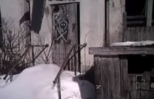 Abandoned building with a door marked by symbols, surrounded by snow and a wooden structure nearby.