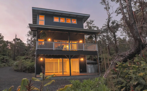 A modern three-story house surrounded by lush greenery, illuminated at dusk with large windows and a balcony.