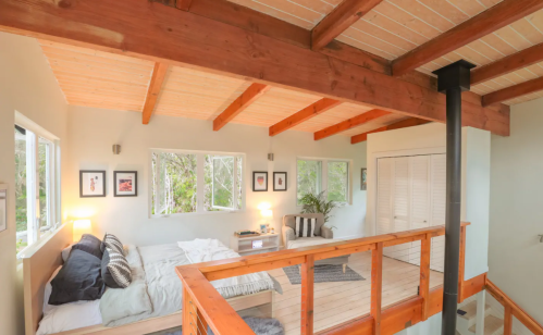 Cozy loft bedroom with wooden beams, large windows, and a small sitting area, surrounded by greenery.