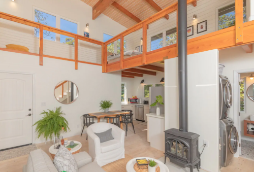 Cozy interior of a modern home with a loft, featuring a living area, dining table, and kitchen with a wood stove.