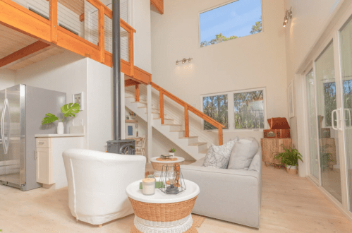 Bright, modern living space with a staircase, large windows, and cozy furniture, surrounded by greenery.