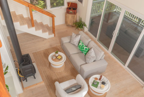A cozy living room with a gray sofa, white chairs, a round coffee table, and a staircase, surrounded by greenery.