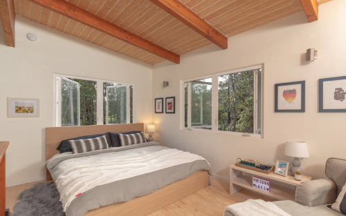Cozy bedroom with a wooden ceiling, large windows, a bed with gray and white bedding, and framed artwork on the walls.