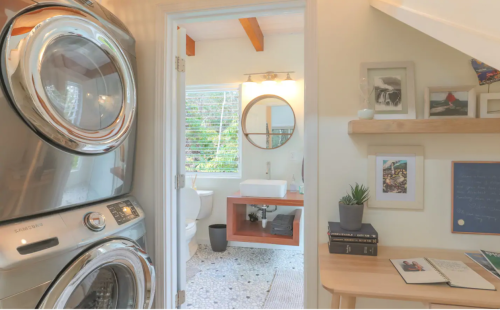 A modern laundry area leads to a bright bathroom with a round mirror, sink, and wooden accents.