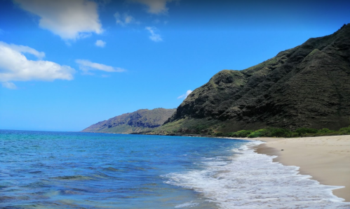 A serene beach with gentle waves, clear blue water, and lush green mountains under a bright blue sky.