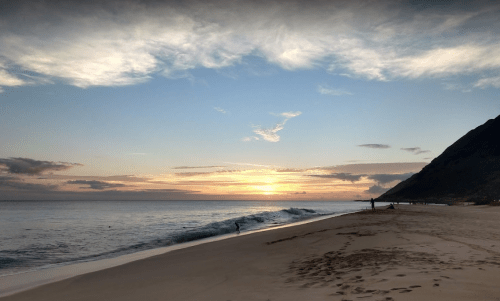 A serene beach at sunset, with gentle waves lapping the shore and a mountain silhouette in the background.