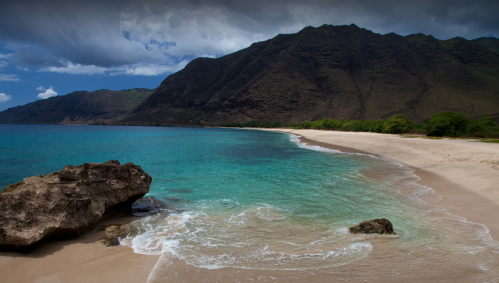 A serene beach with turquoise water, soft sand, and a backdrop of lush mountains under a partly cloudy sky.