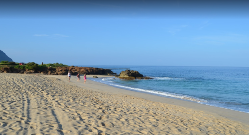 Two people walk along a sandy beach beside calm blue waters and rocky formations under a clear sky.