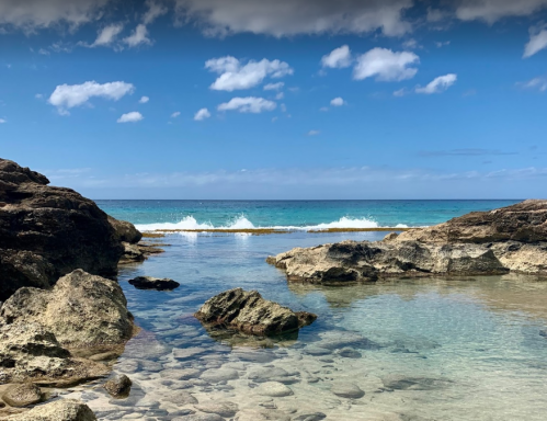 A serene beach scene with clear water, rocky formations, and gentle waves under a blue sky with scattered clouds.