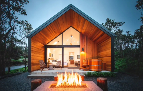 A modern wooden cabin with large windows, a fire pit in front, and surrounded by greenery. Cozy and inviting atmosphere.