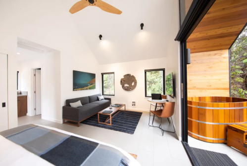 Modern room with a sofa, desk, and large windows, featuring a wooden hot tub on a balcony. Bright and airy design.