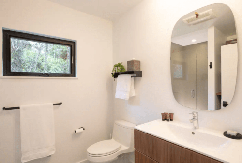 Modern bathroom featuring a toilet, sink with a mirror, and a window with greenery outside. White towels hang on a rack.